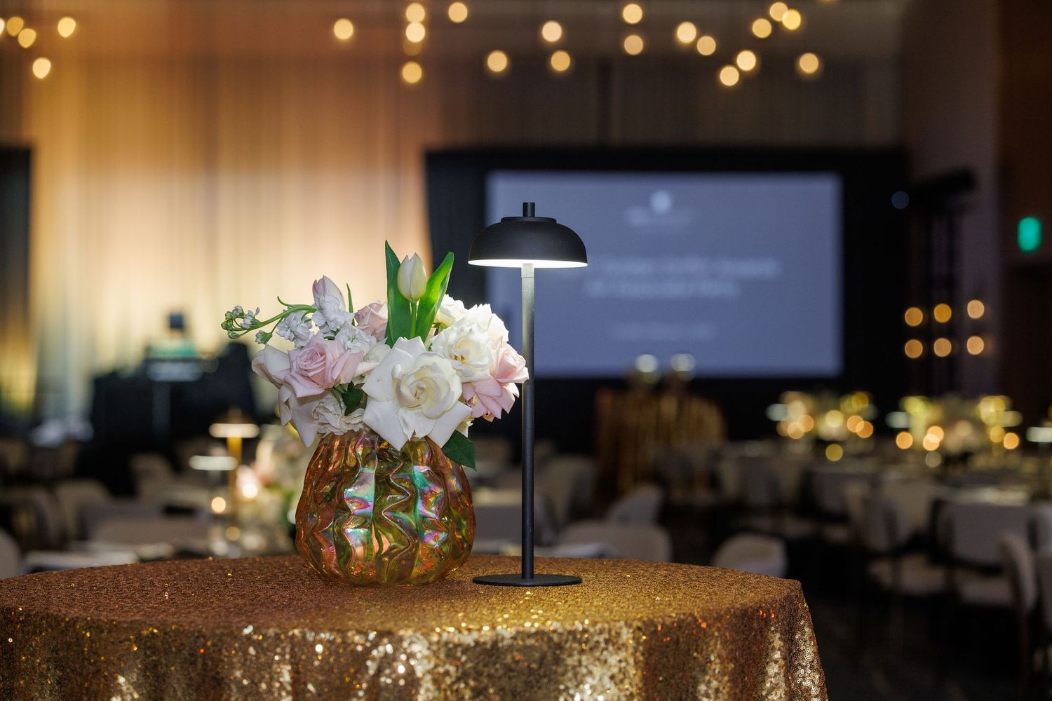 Floral centerpiece on gold sequin tablecloth with blurred event space and bokeh lights in background.