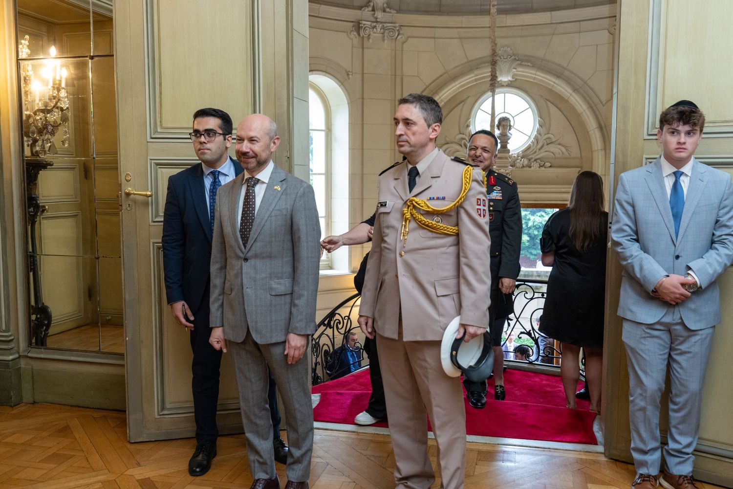 People in formal attire gather in an elegant room with ornate walls and chandelier lighting.