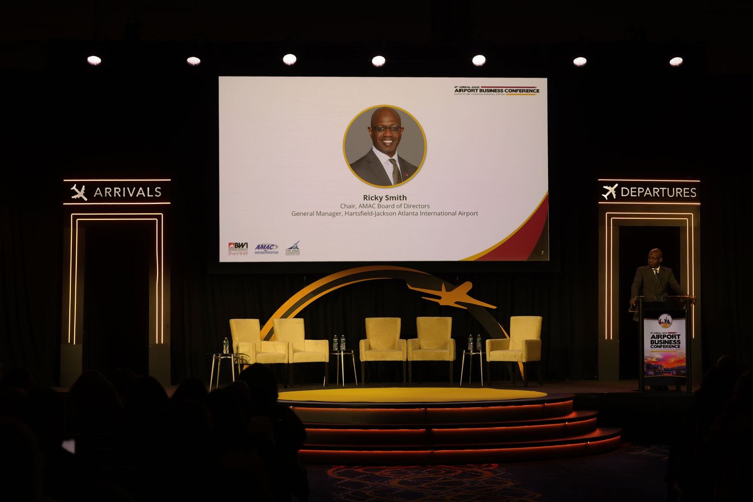 Speaker presentation at an airport-themed conference stage with arrivals and departures signs and a profile photo on screen.