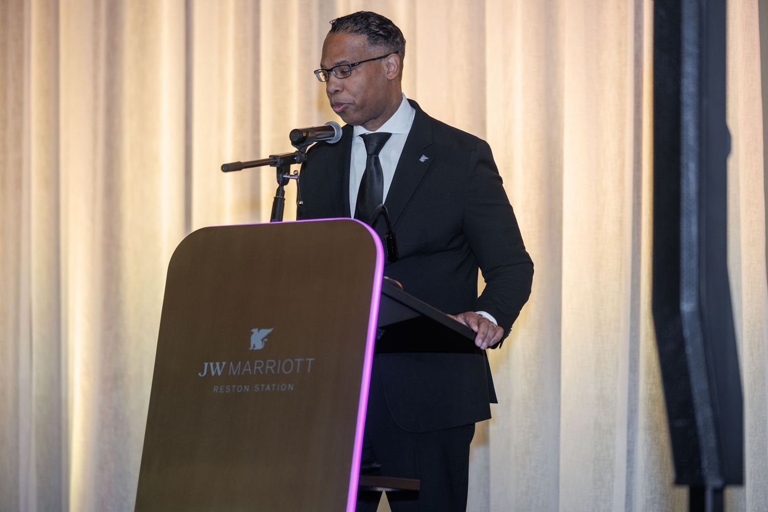 Man in suit speaking at JW Marriott podium during formal event.