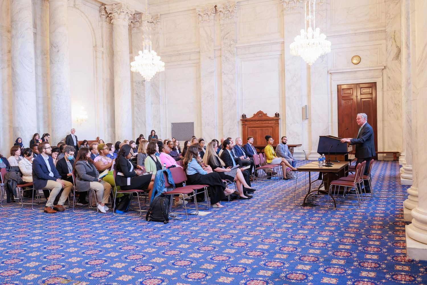People seated in rows on blue patterned carpet in an elegant white room with crystal chandeliers.