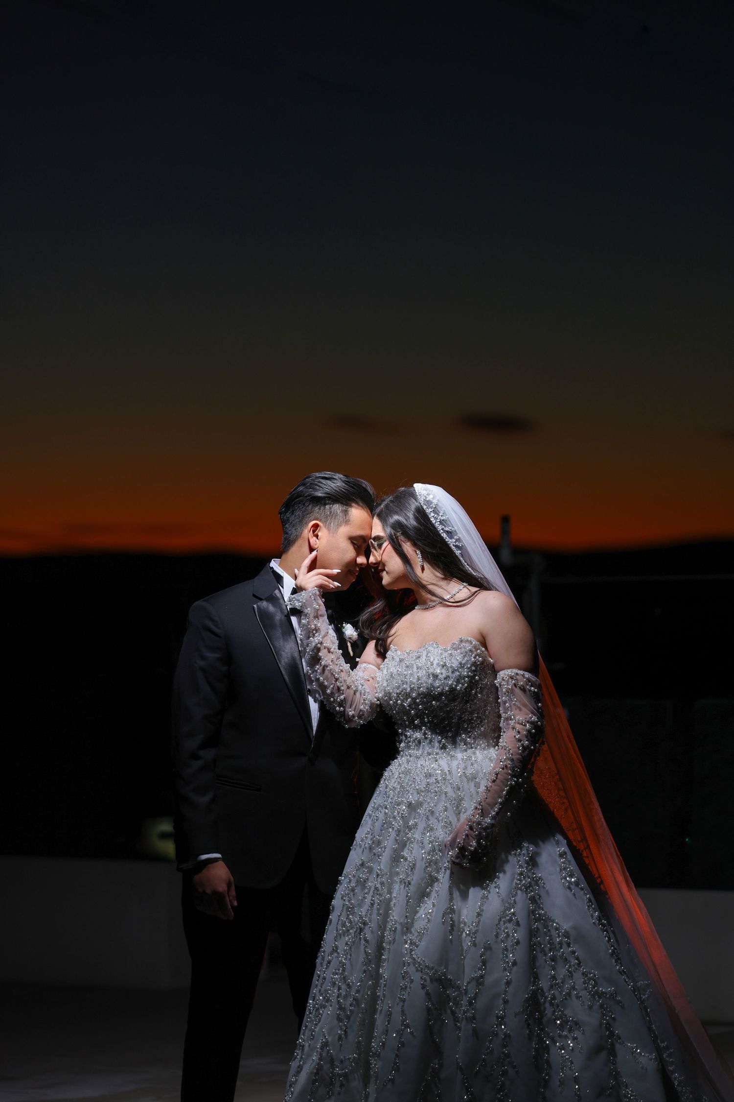 Bride and groom share romantic sunset moment, she in beaded gown with veil, he in black tuxedo, against twilight sky.