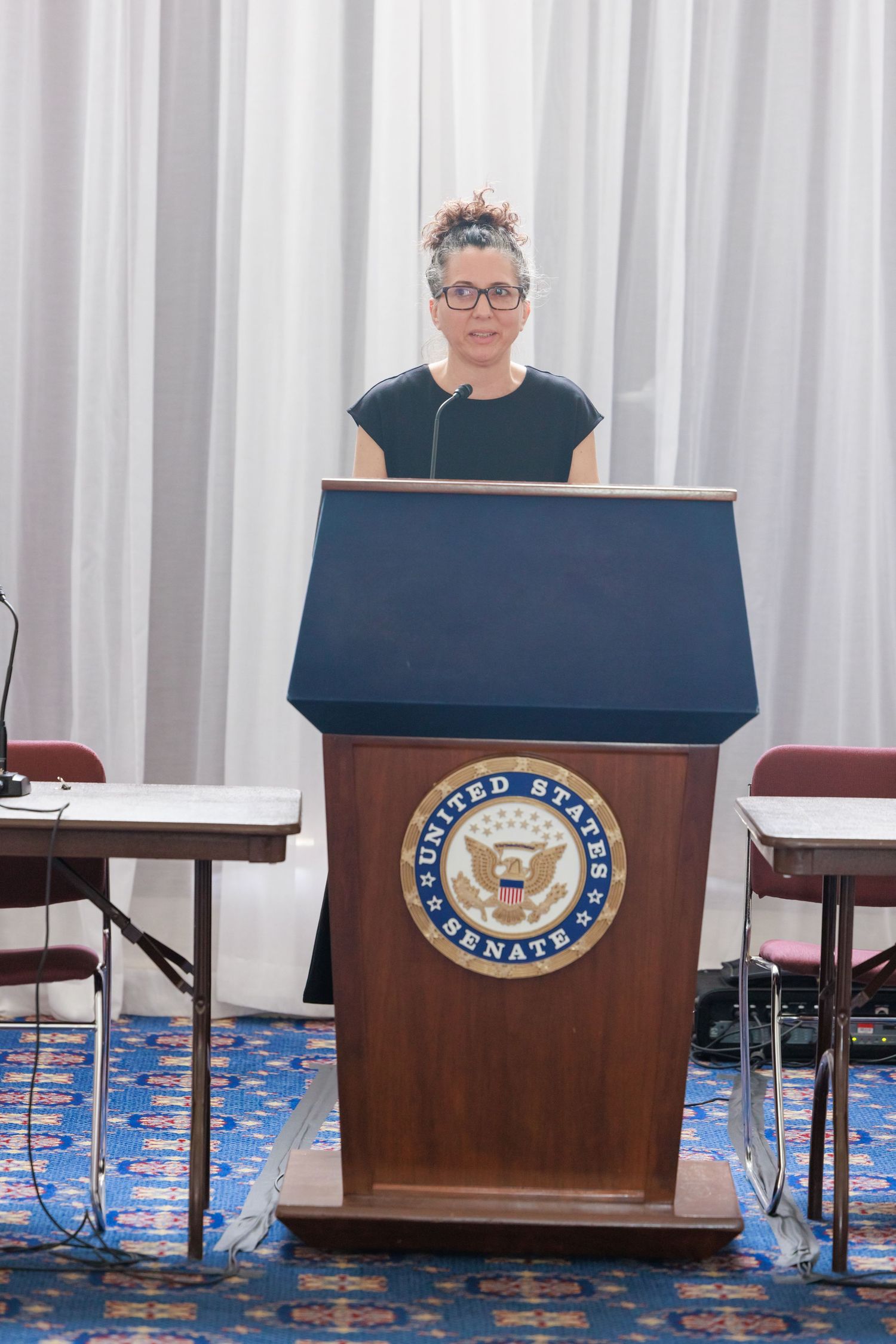 Empty podiums with Senate seal stand in a row against white curtains in a government hearing room.