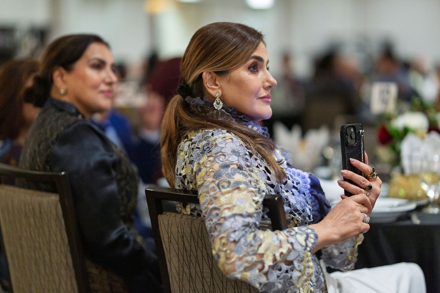 Two people seated at a formal event, with one holding a mobile phone while wearing an ornate gold and silver patterned dress.