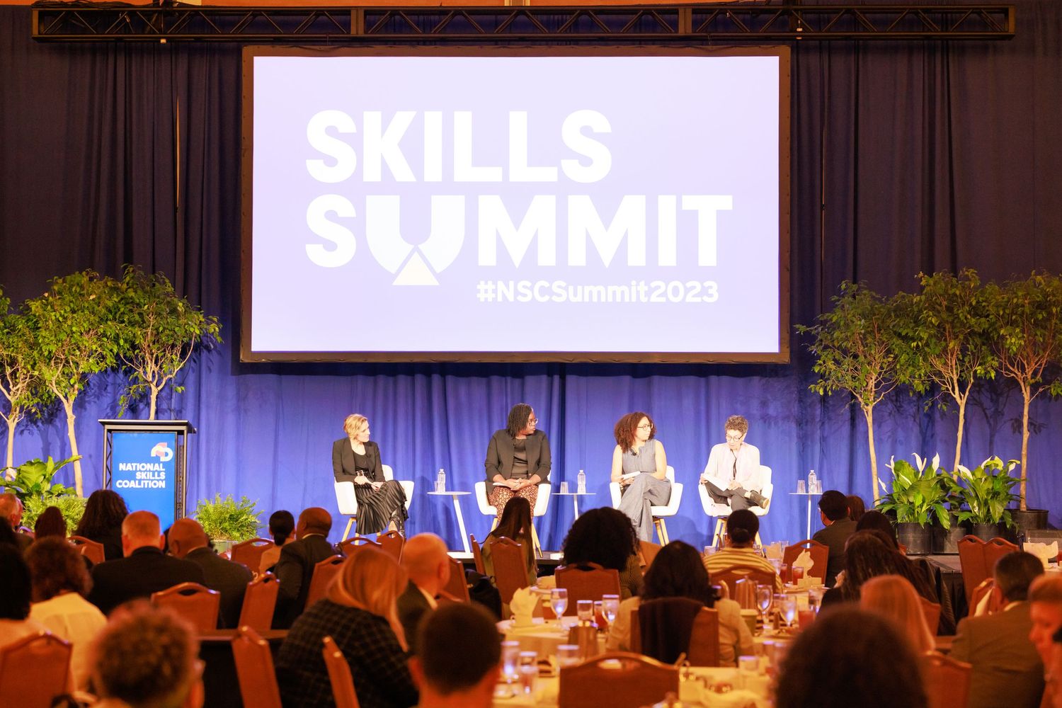 Business conference panel discussion on stage with audience members seated in a darkened auditorium.