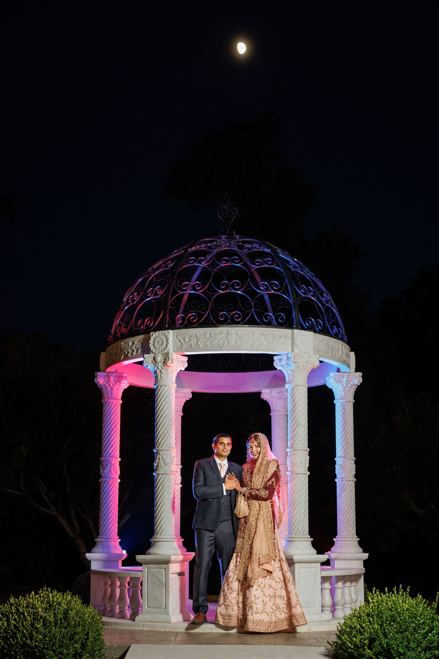 Illuminated white stone gazebo with pink and blue lighting creates romantic ambiance during nighttime wedding ceremony.