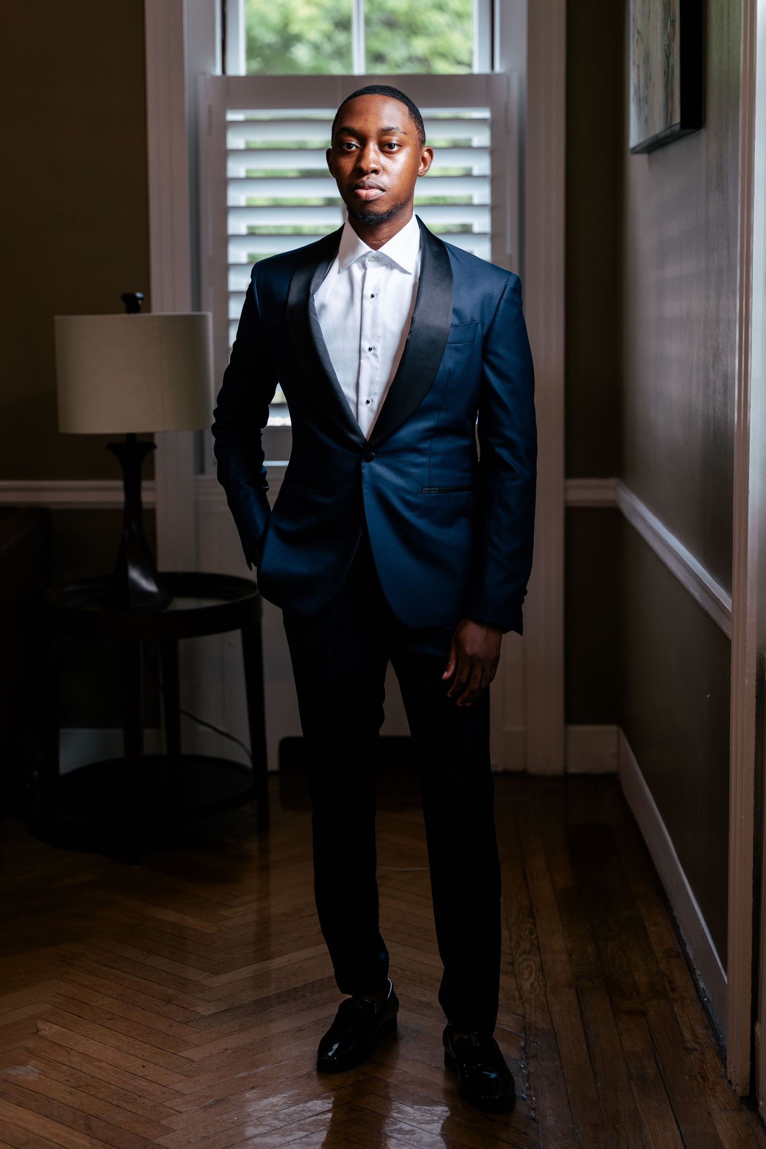 A person in a formal navy blue tuxedo and bow tie stands confidently in a hallway with hardwood floors and window light.