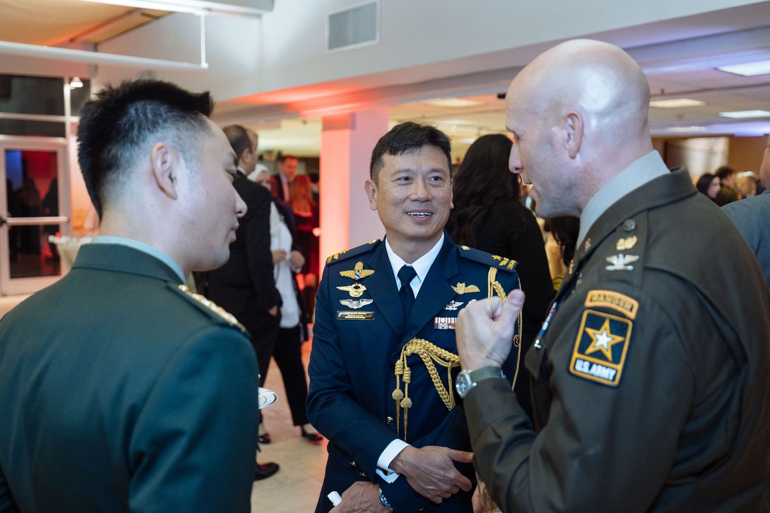 Military personnel in formal uniforms gather at an indoor event with red and blue lighting.