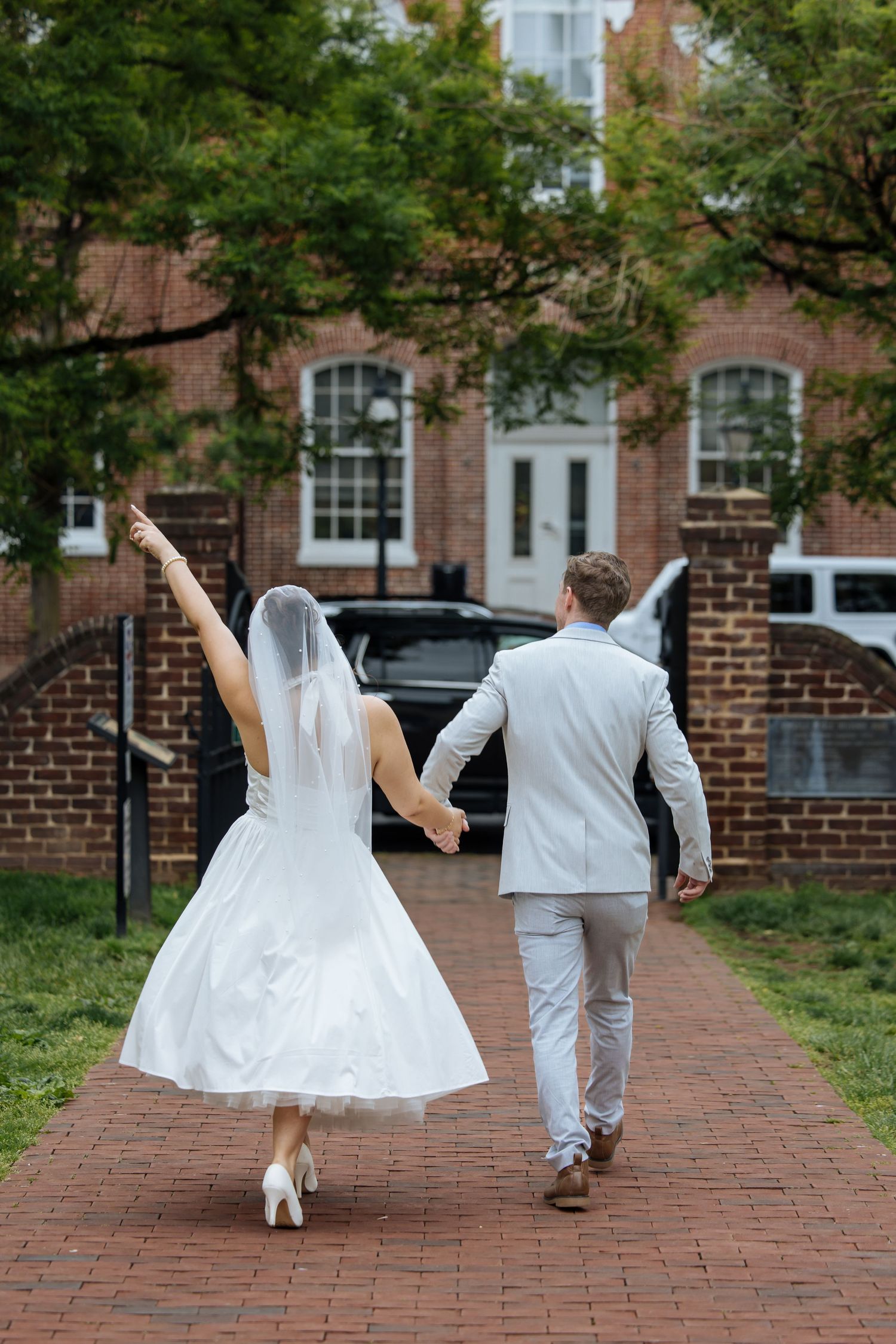A bride and groom walk away on a brick path while holding hands and raising their arms in celebration.