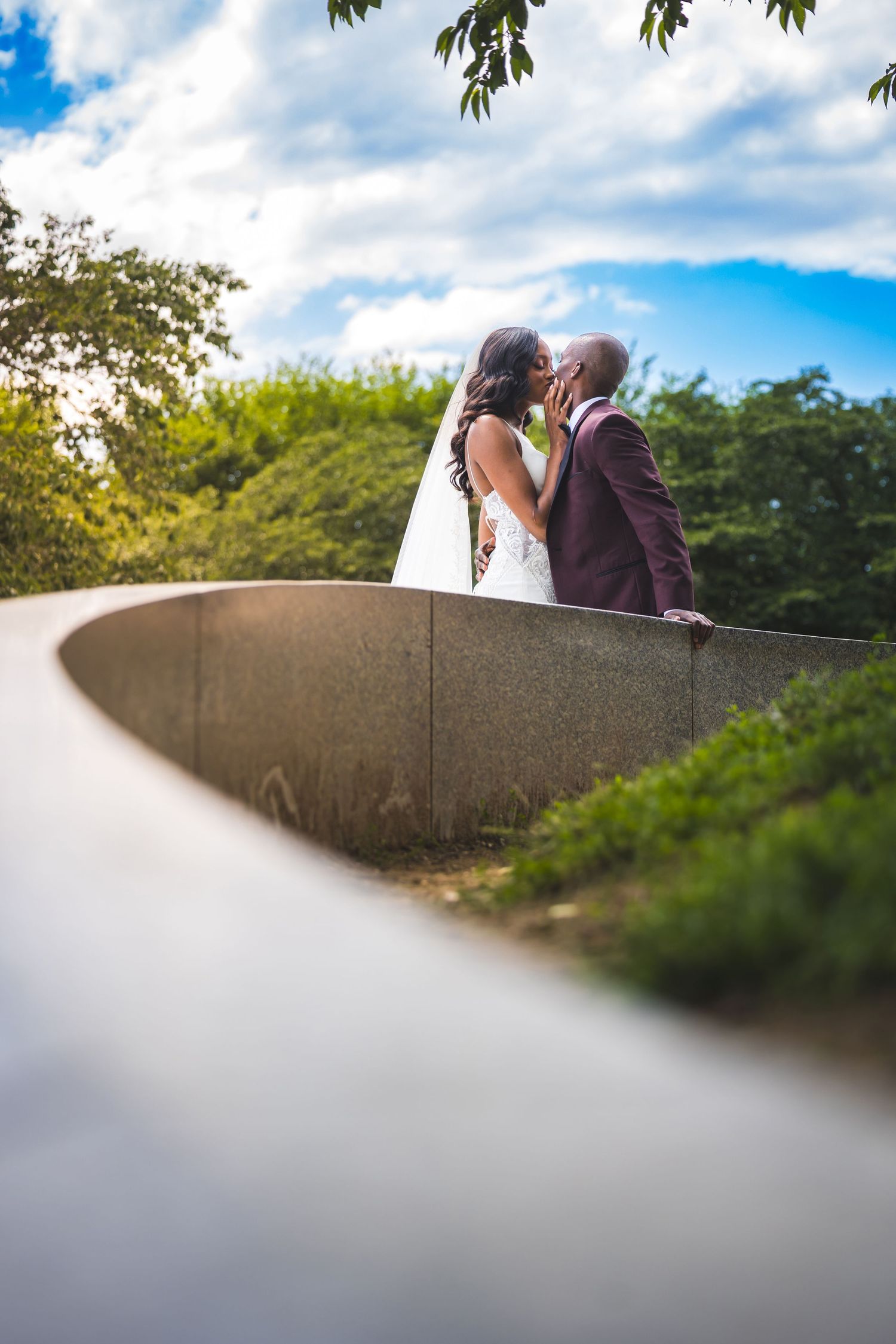 A couple shares a tender moment while sitting atop a curved concrete wall surrounded by green foliage and blue sky.