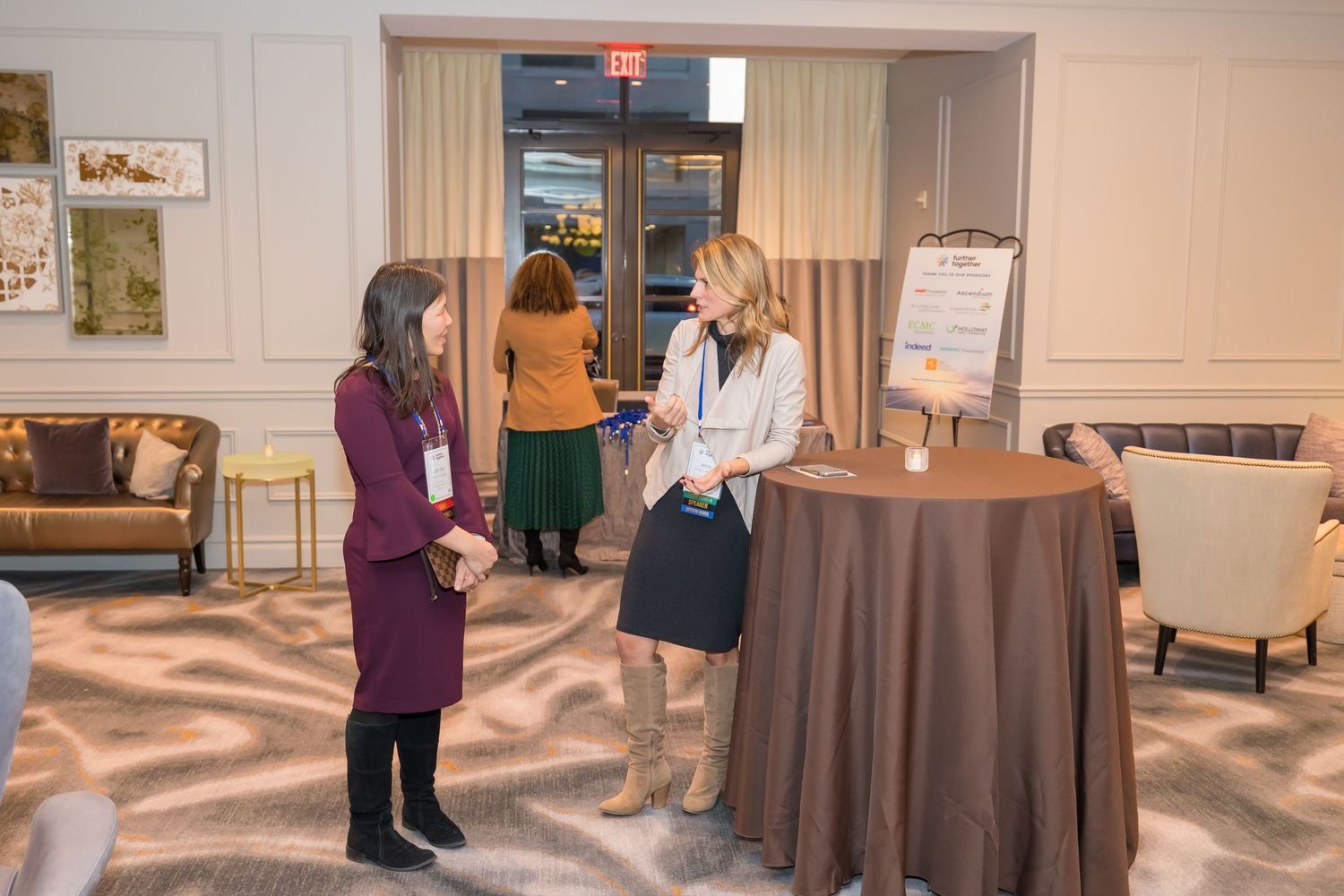 Two people stand and converse in a carpeted event space with brown tablecloths and beige walls.