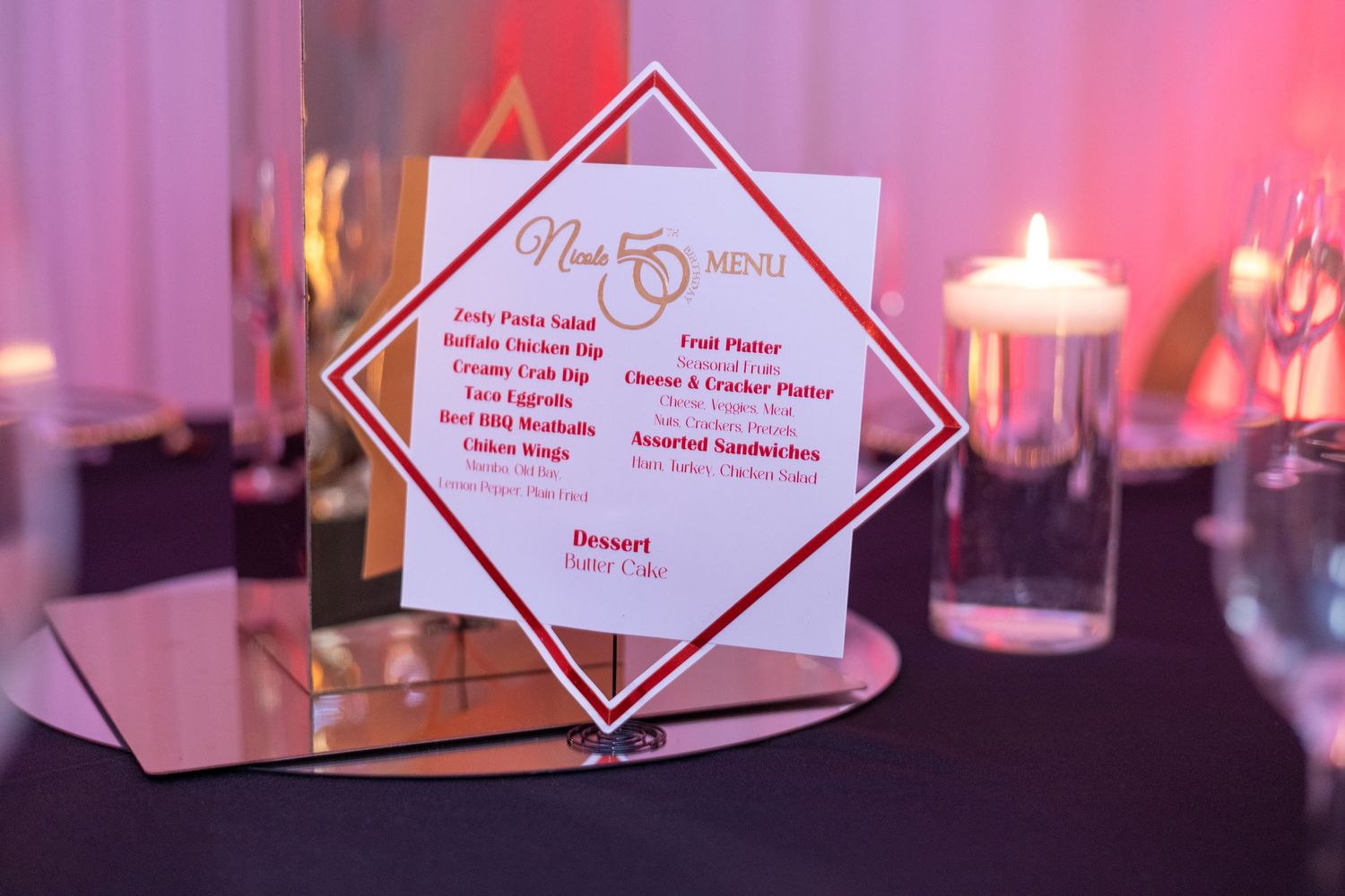 Elegant wedding reception menu card displayed on a mirrored stand with candles and pink lighting in the background.