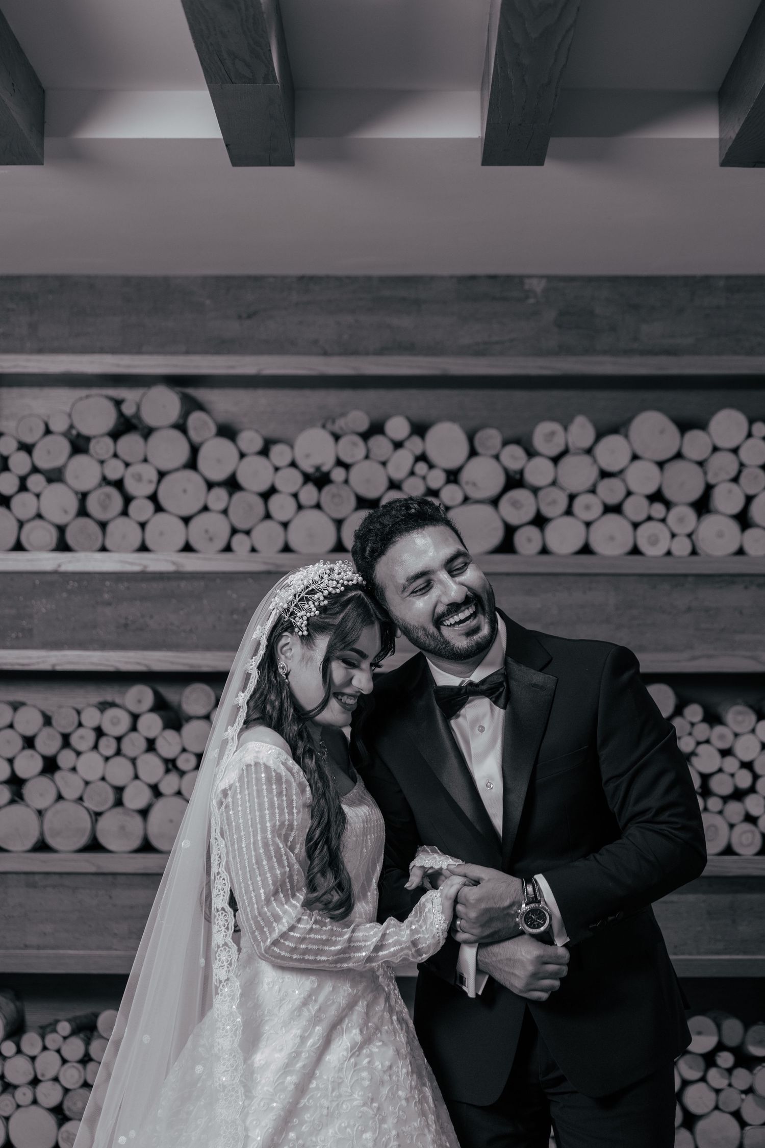 Black and white wedding photo of a couple sharing a joyful moment in front of a rustic wood wall with stacked logs.