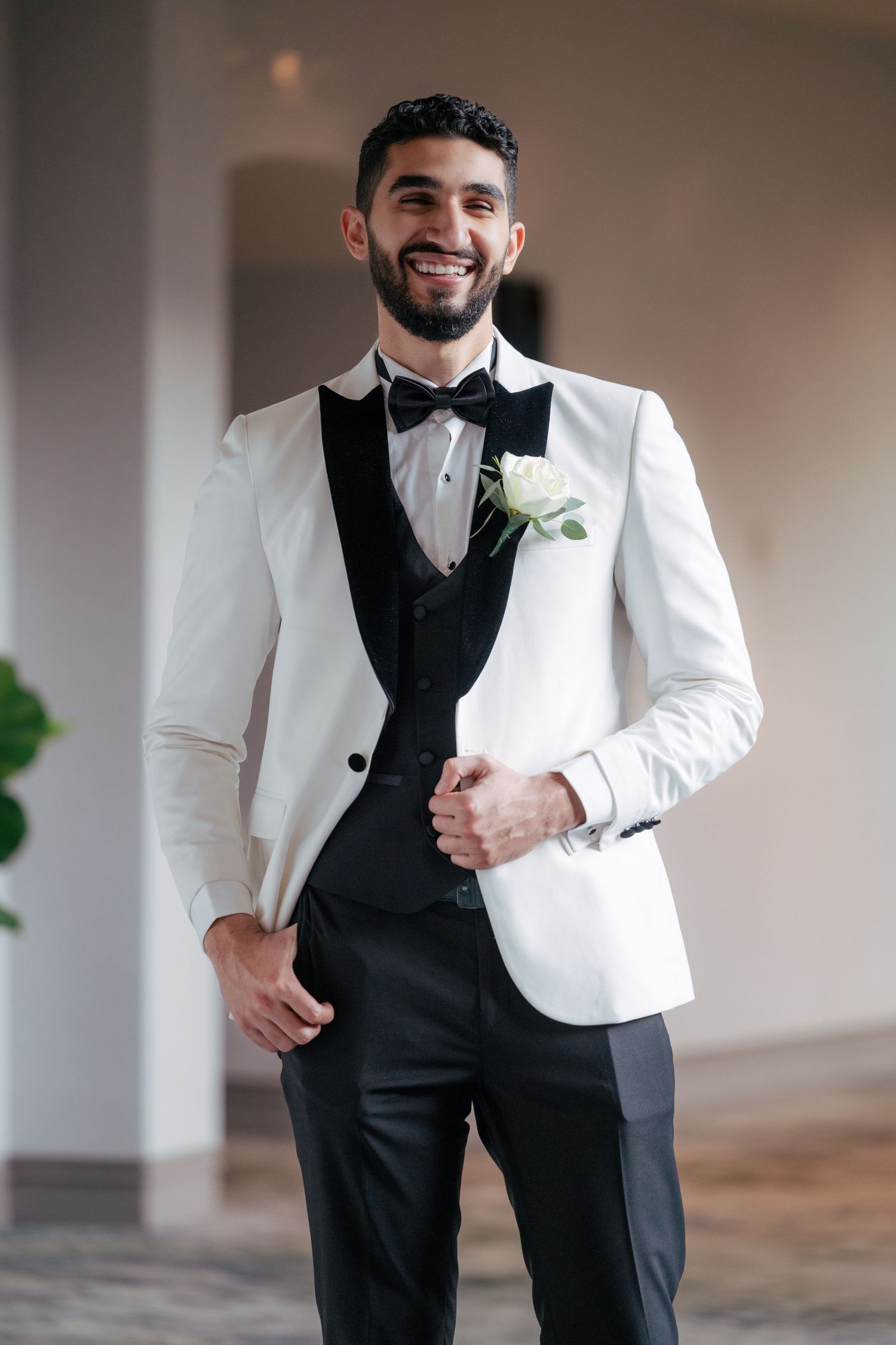A groom in a classic black and white tuxedo smiles while adjusting his jacket in an elegant indoor setting.