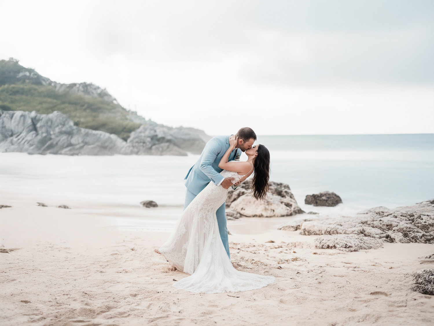 Romantic coastal wedding photography session on a scenic beach with rocky cliffs and cloudy sky in the background.