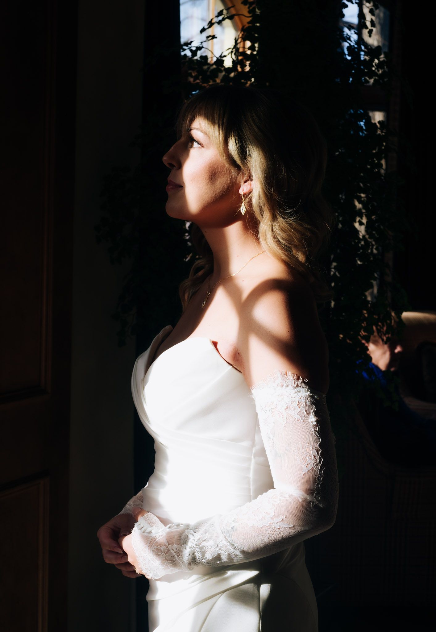 A bride in a white long-sleeved wedding dress stands in dramatic side lighting creating shadows and highlights.