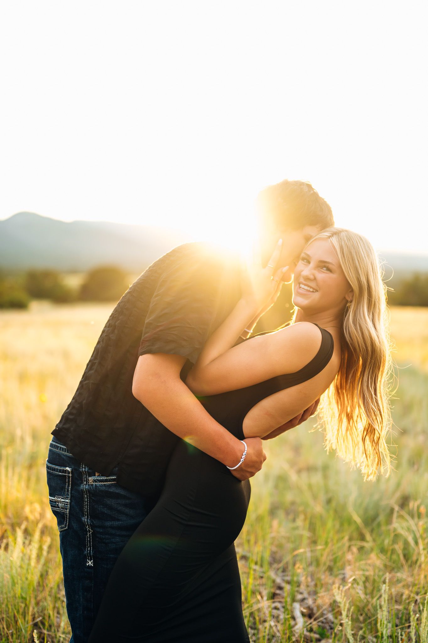 A couple embraces in a sun-drenched meadow during golden hour, backlit by warm sunlight in a romantic outdoor setting.
