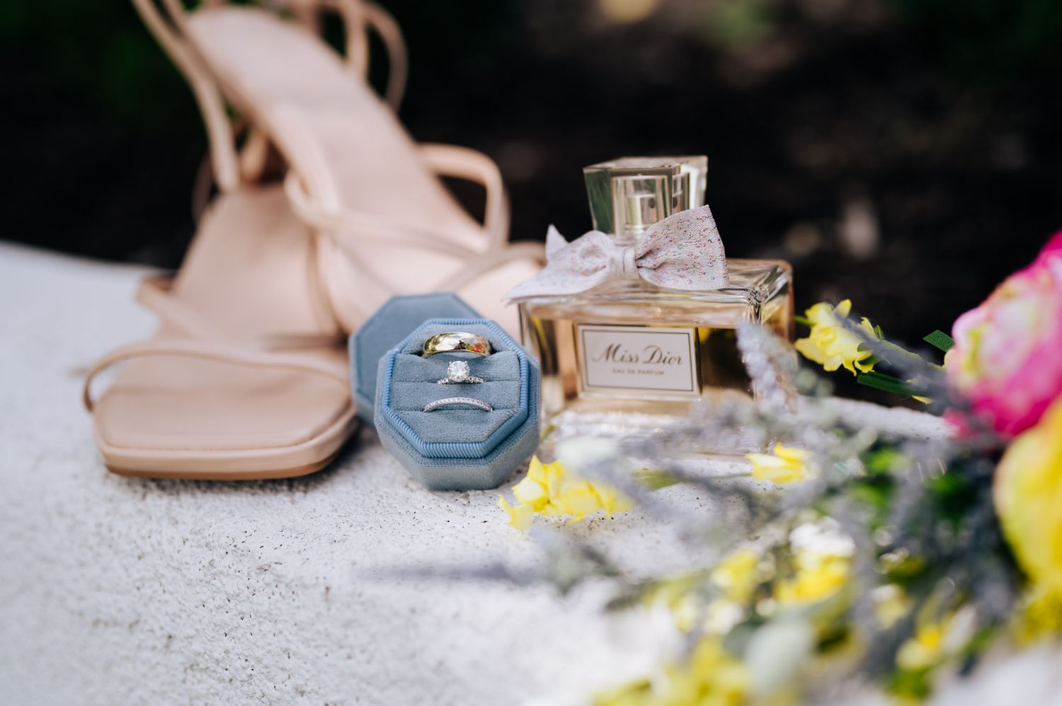 Elegant wedding accessories including perfume bottle, pink shoes, and delicate yellow flowers arranged on white surface.
