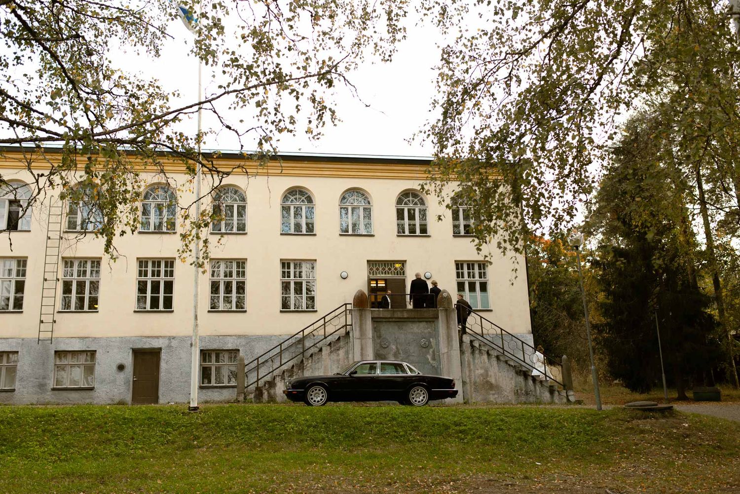 Vaaleankeltainen korkea kivirakennus, jonka portailla juhlavieraita ja edustalla hääauto.