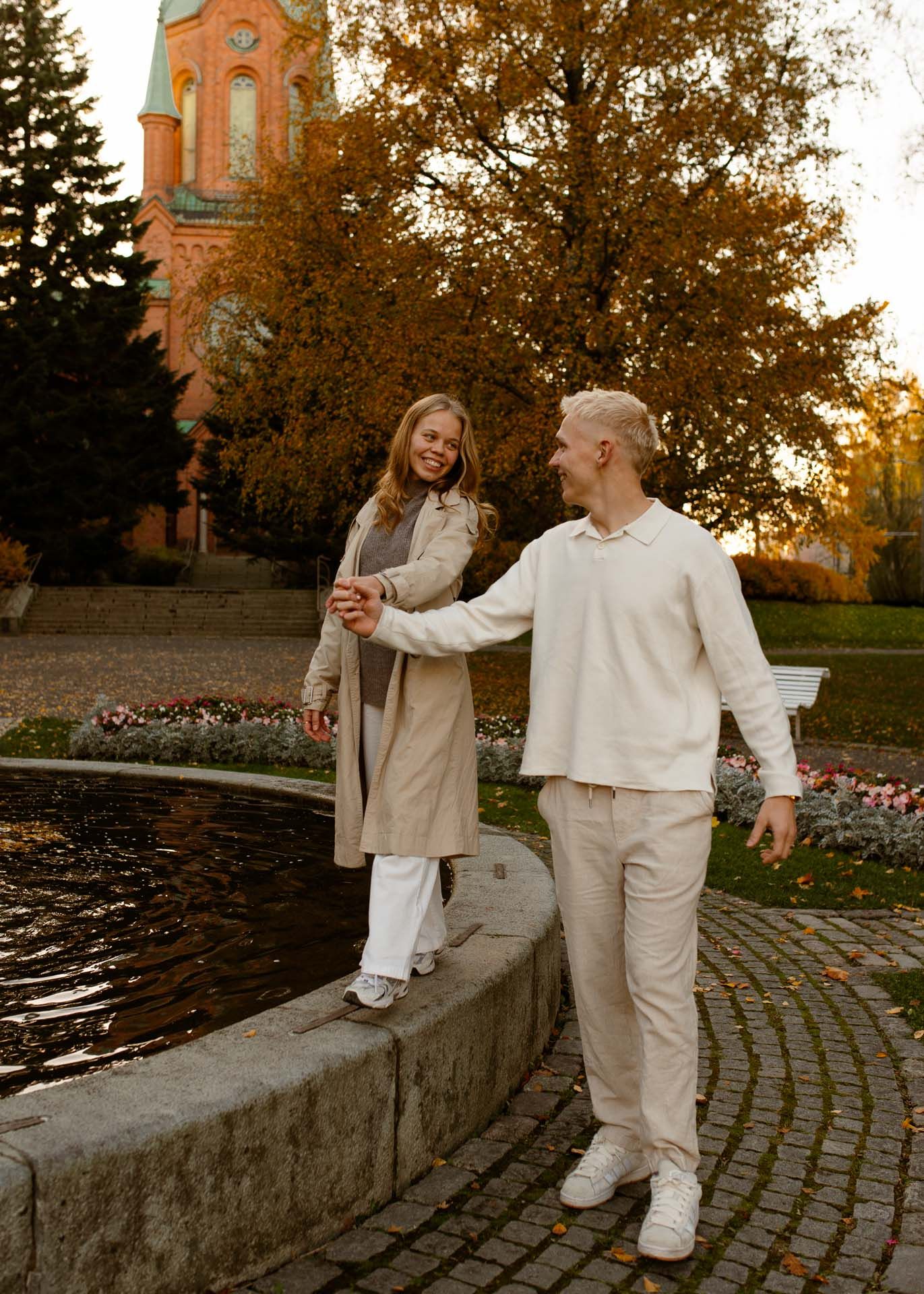 Tyttö kävelee suihkulähteen reunalla pojan pitäessä häntä kädessä.