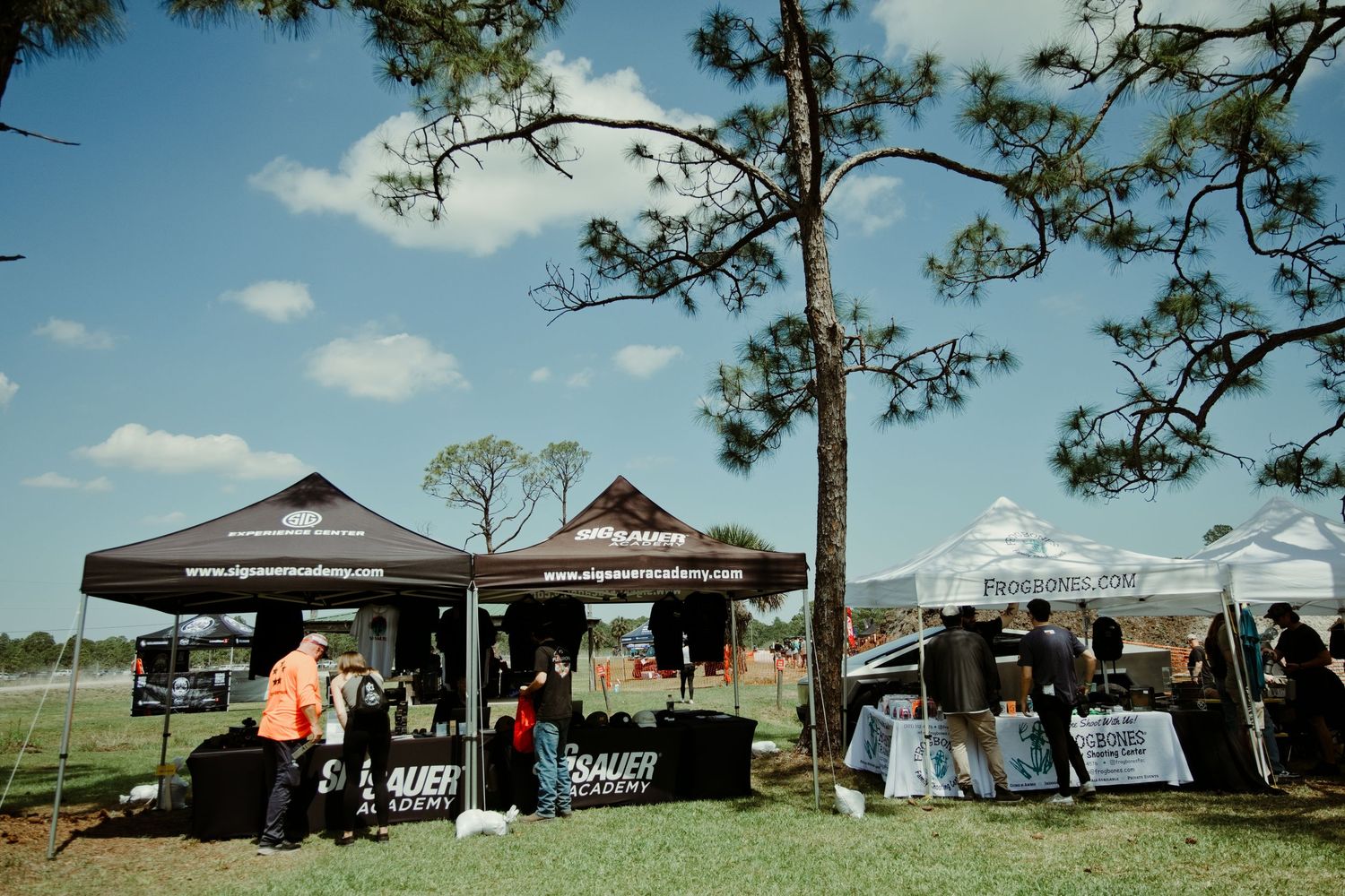 Outdoor vendor booths with dark canopies at a shooting sports event set among tall pine trees on a sunny day.