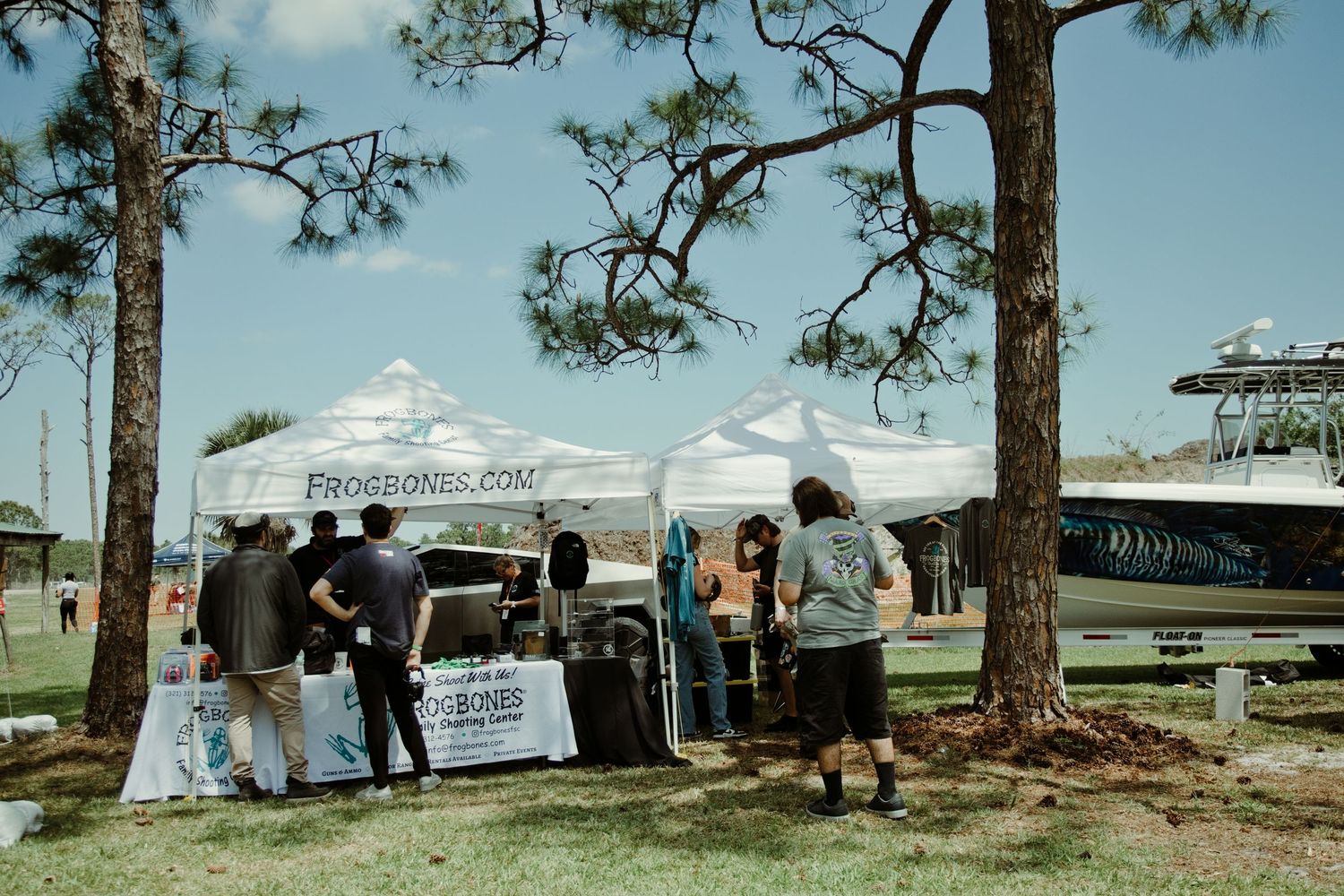 Outdoor vendor booths at ProsBones.com event near waterfront with pine trees and a boat visible in background.