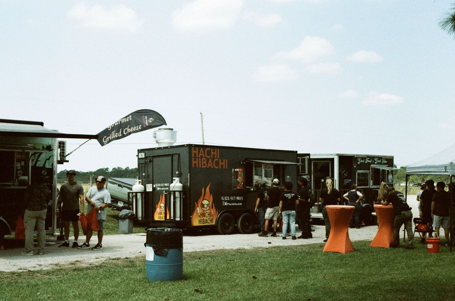 Outdoor food truck festival with Hachi Hibachi truck and vendors on a sunny day, people gathered on green grass.