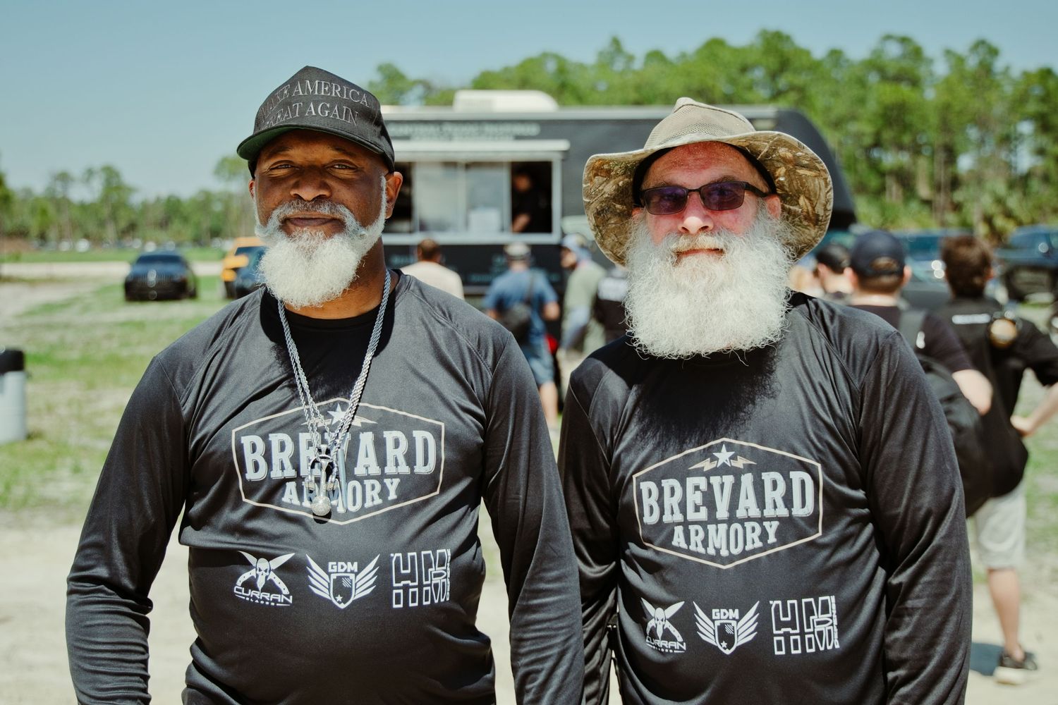 Two men with white beards wearing Brevard Armory black long-sleeve shirts pose together at an outdoor event.
