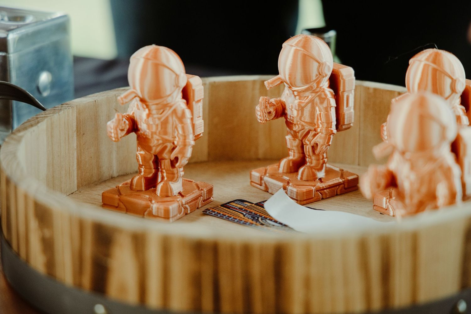 Small orange 3D printed astronaut figurines displayed in a wooden bowl at an event.