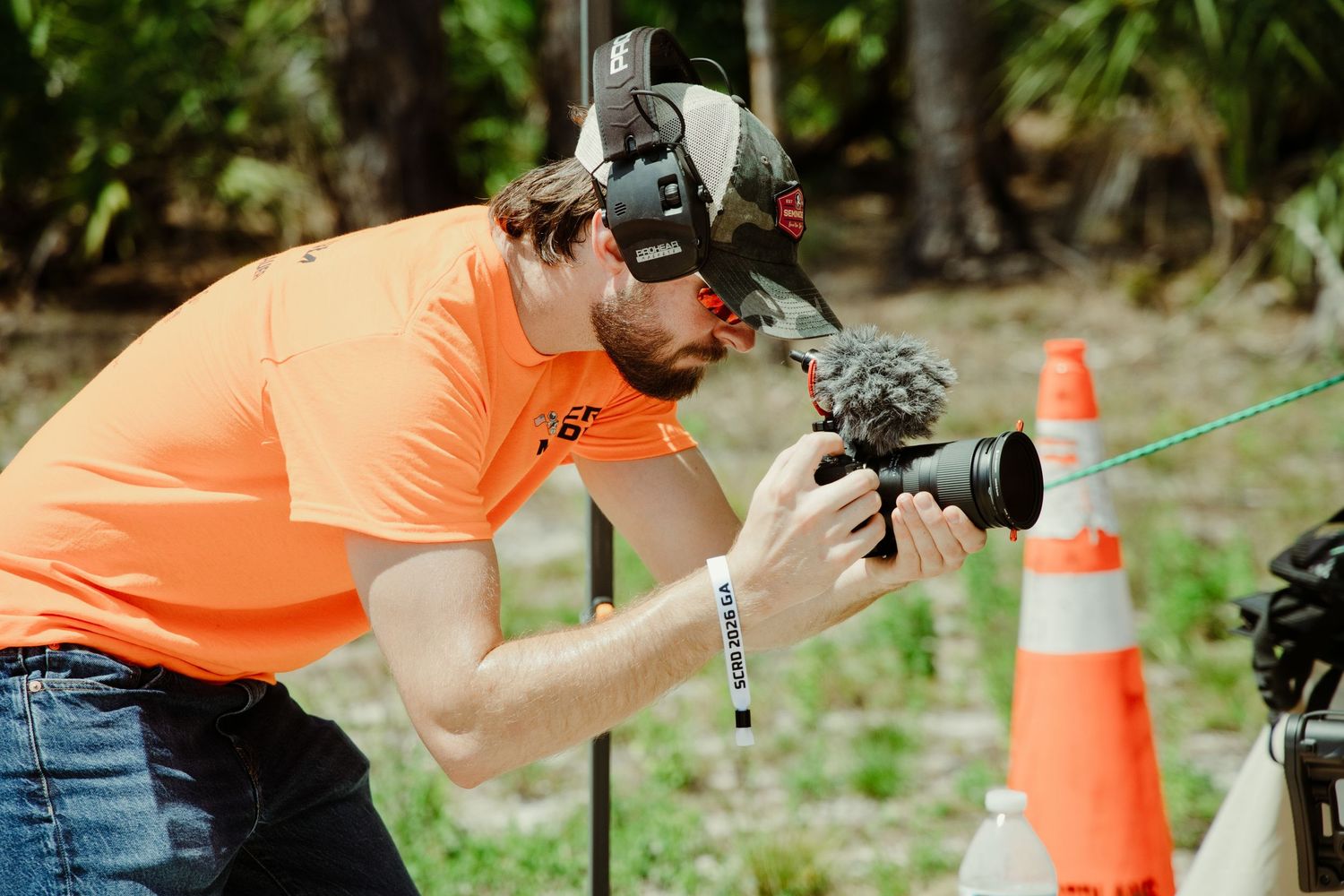 Photographer with boom mic and camera filming outdoors near orange traffic cones at shooting event.