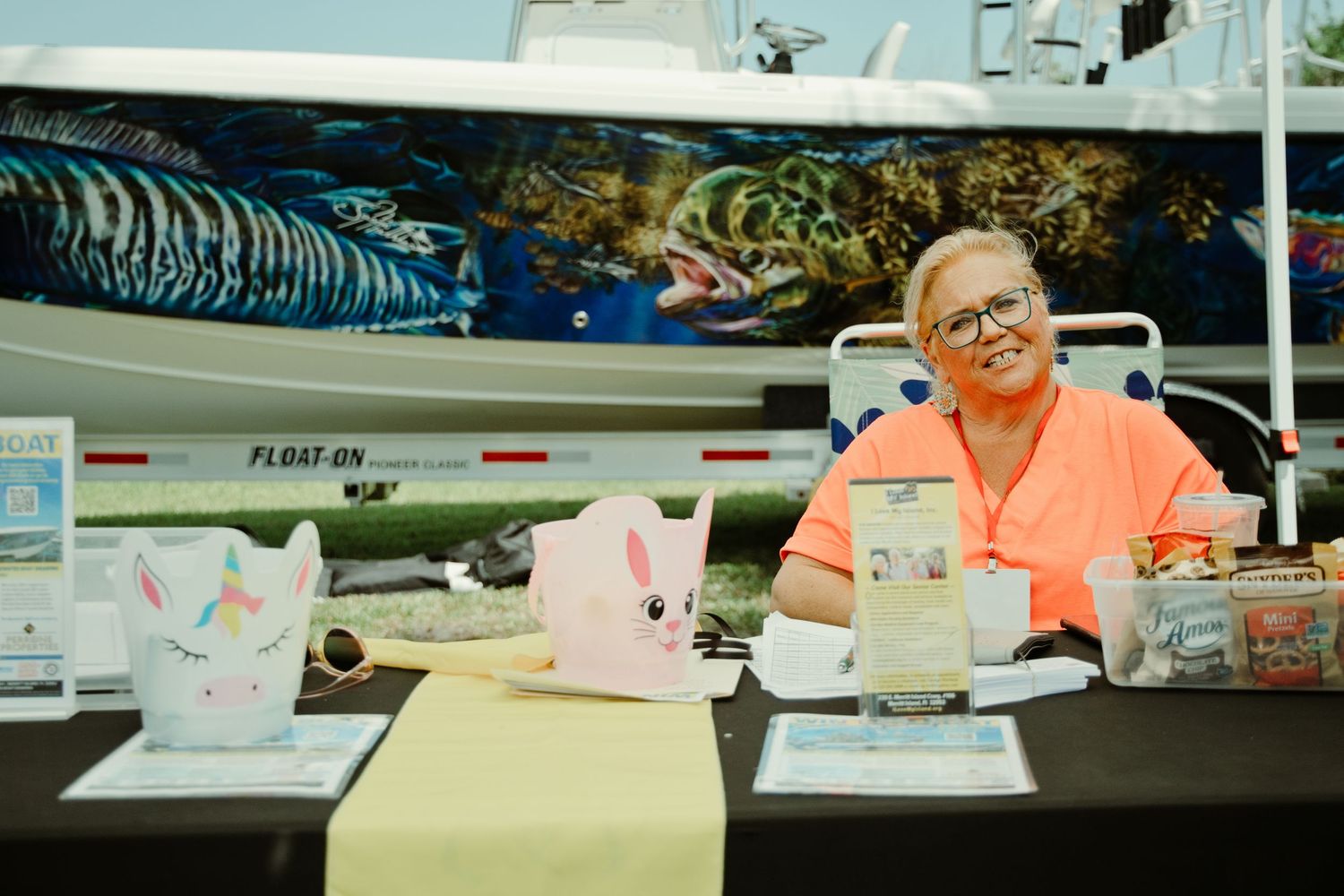 Woman smiling at vendor booth with colorful fishing boat mural backdrop at outdoor event.