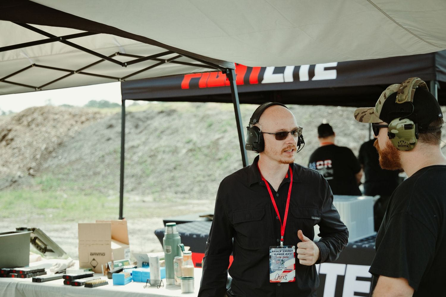 Man with headphones and lanyard stands at outdoor shooting range event booth under a black canopy tent.