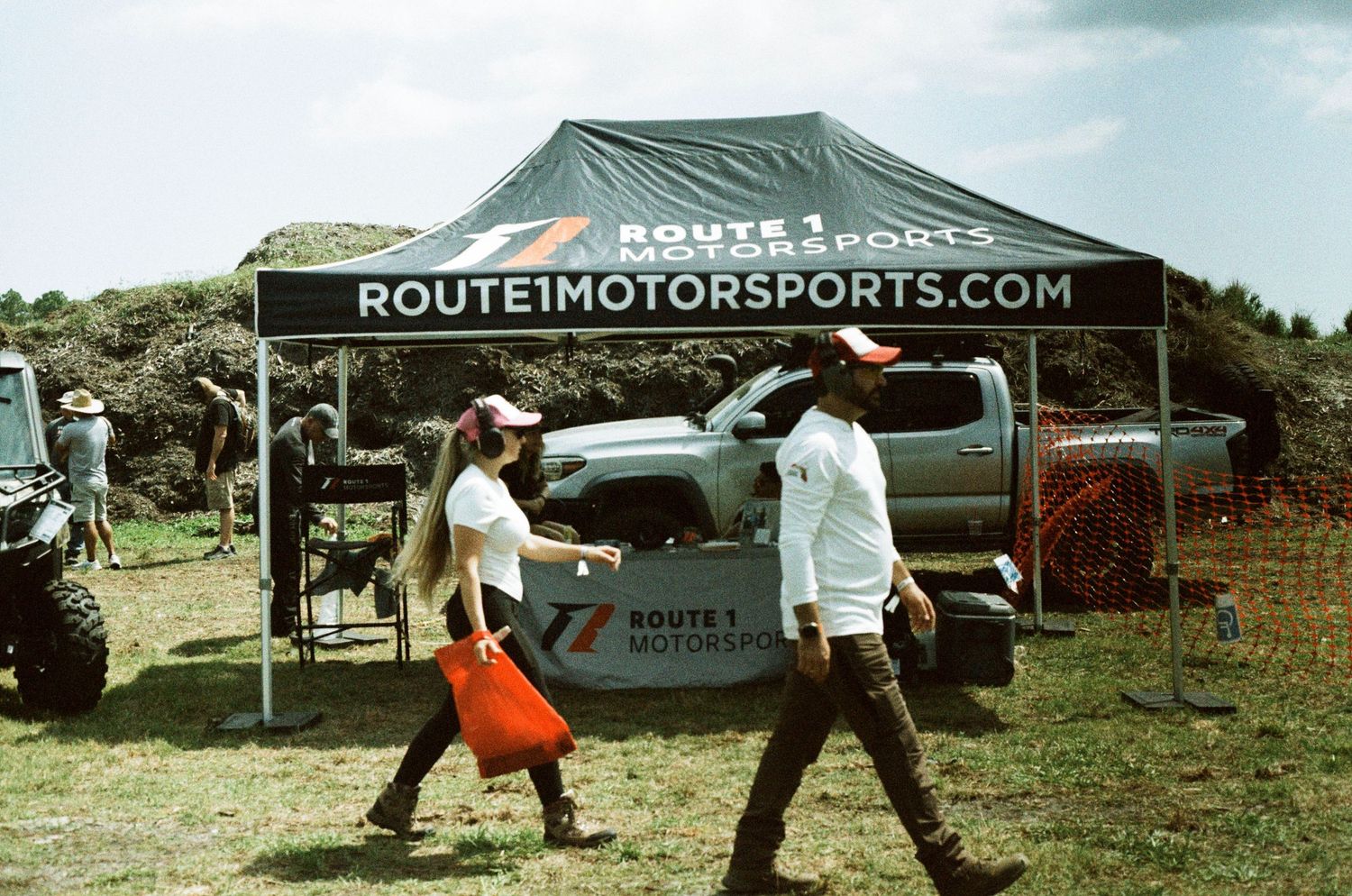 Two attendees walk past a Route 1 Motorsports branded tent at an outdoor motorsports expo event.
