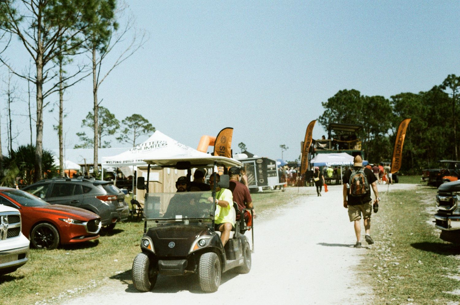 Golf cart and visitors navigate a sunny outdoor festival grounds lined with vendor tents.