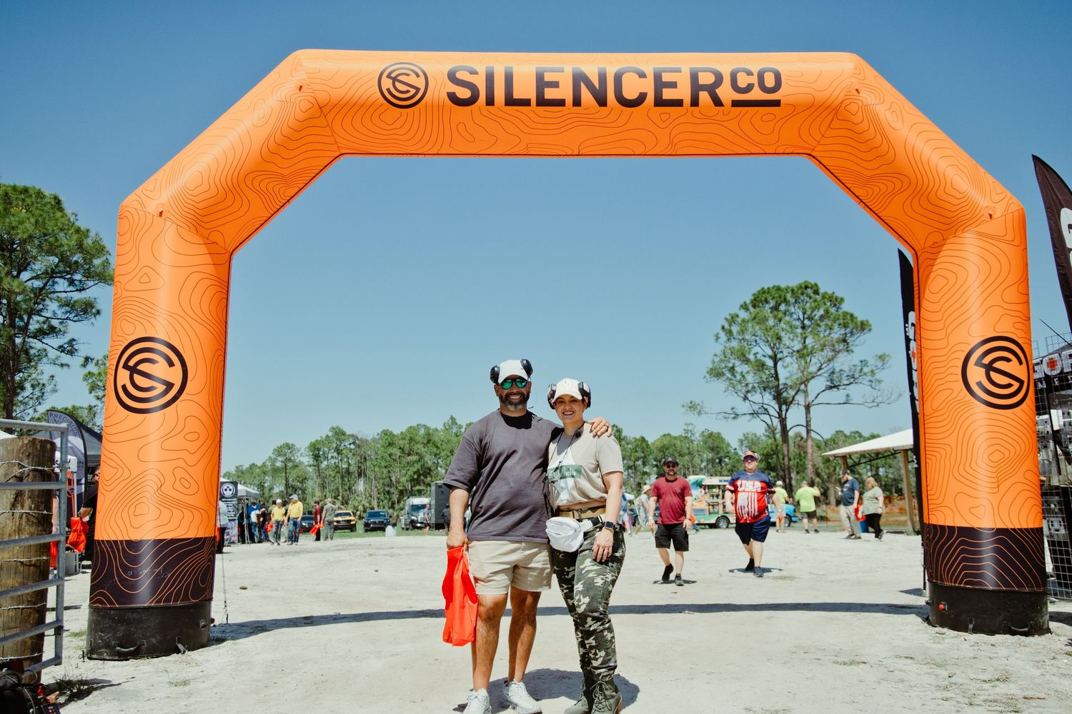 Two people posing under an orange SilencerCo inflatable arch at an outdoor gun expo event.
