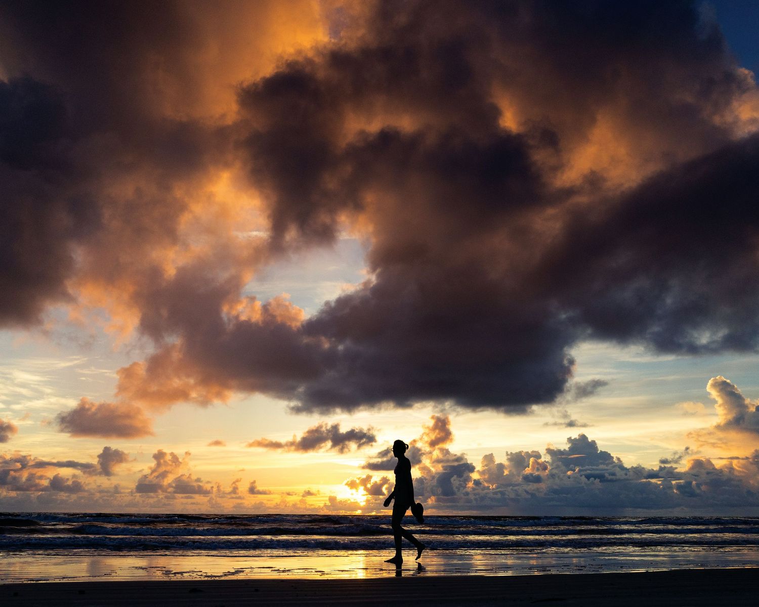 Silhouette of person walking on beach during dramatic orange and purple sunset.
