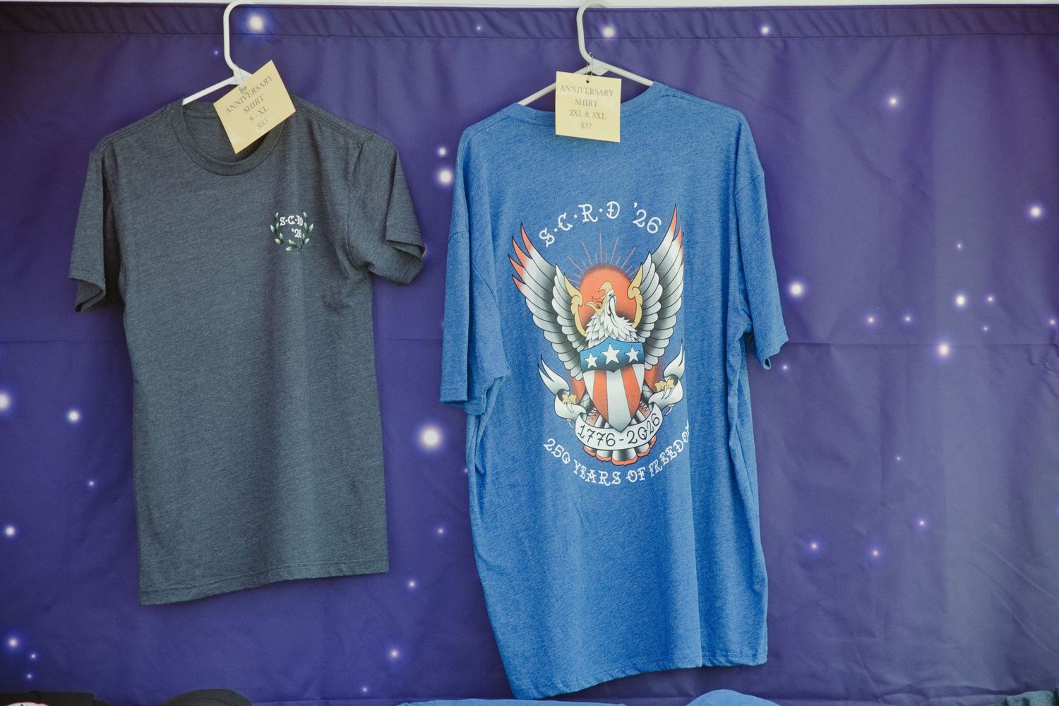 Two blue graphic t-shirts hanging on display against a starry purple backdrop at an outdoor event.
