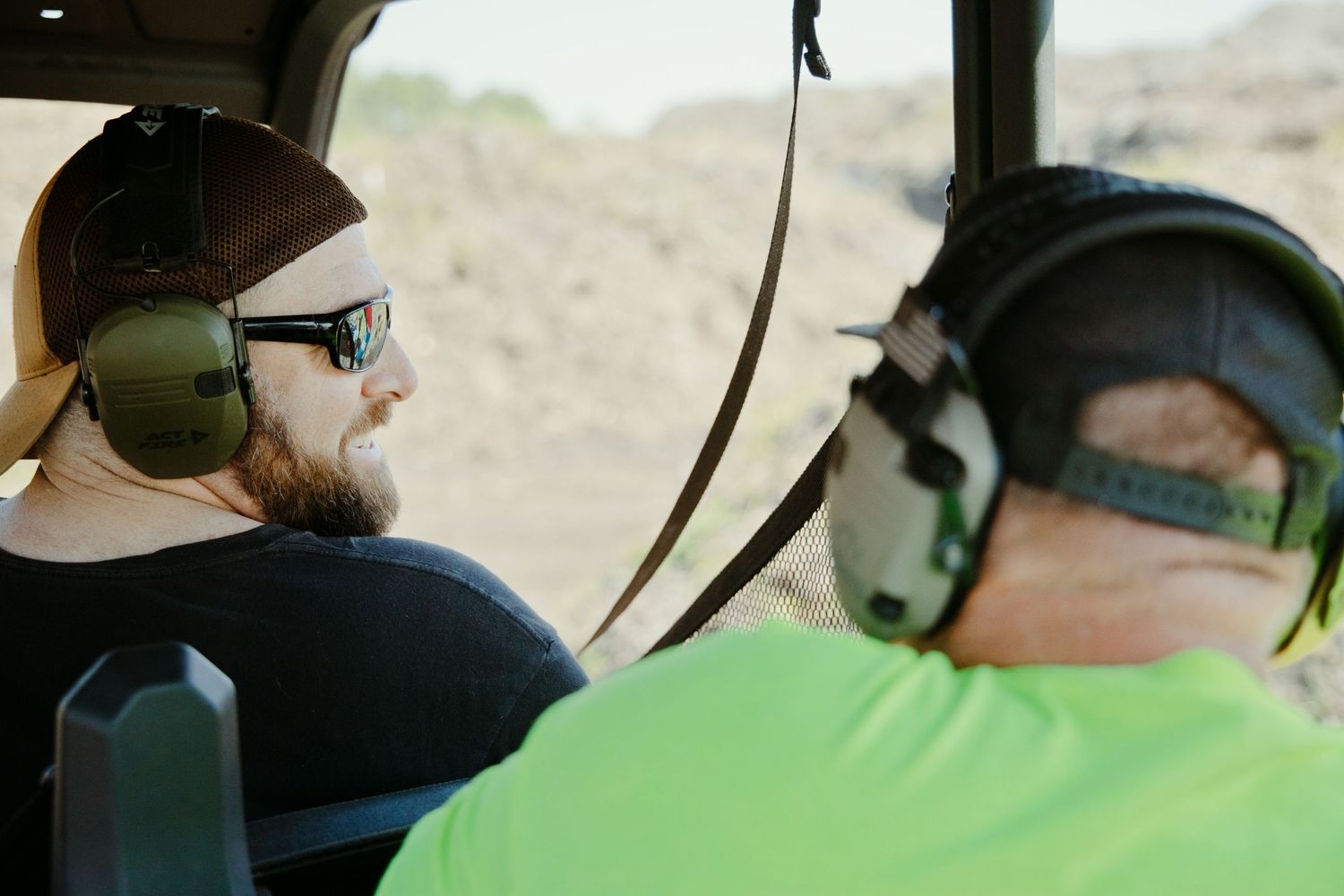 Two men wearing ear protection riding in a vehicle at an outdoor shooting range event.