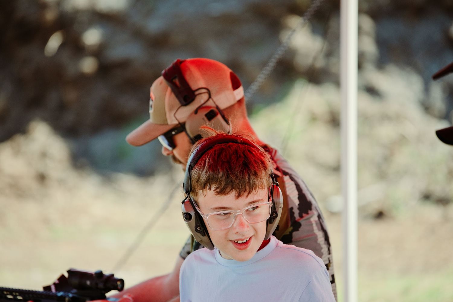 Young boy wearing glasses and headphones at an outdoor shooting range with adult supervisor behind him.