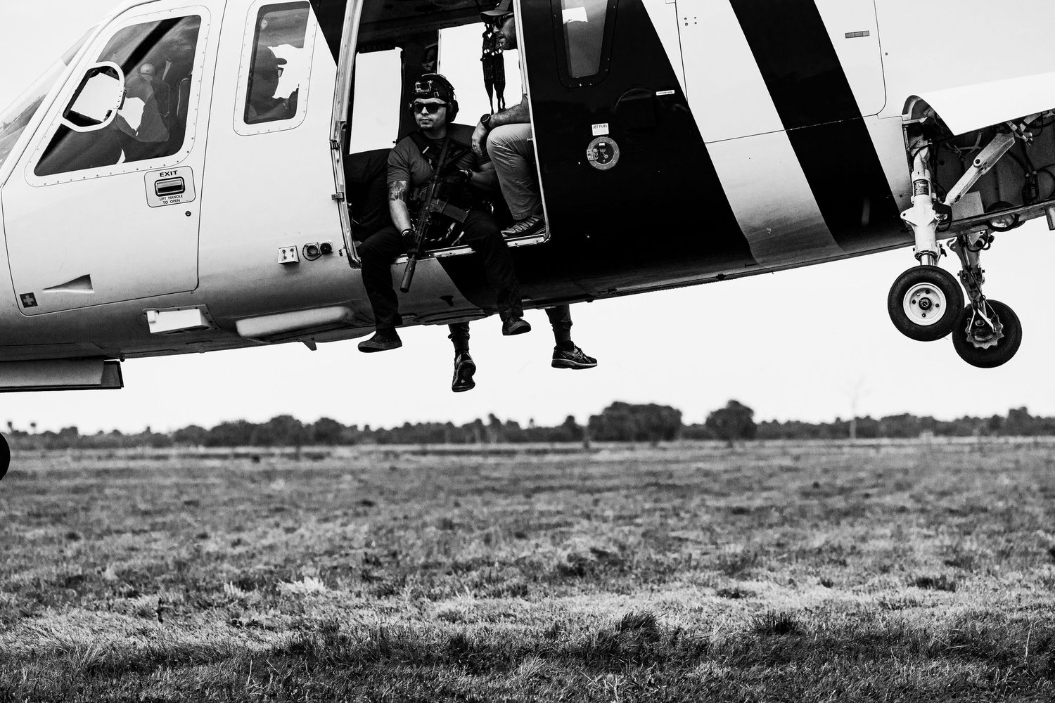 Military helicopter landing on grassy field with personnel nearby in black and white.