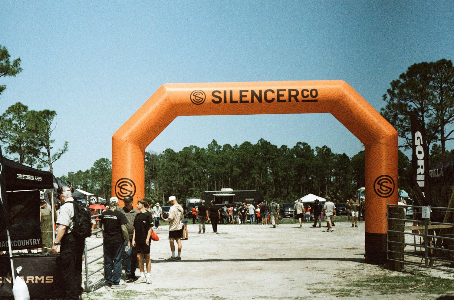 Large orange SilencerCo inflatable arch gate at an outdoor shooting sports event with attendees and vendor booths.
