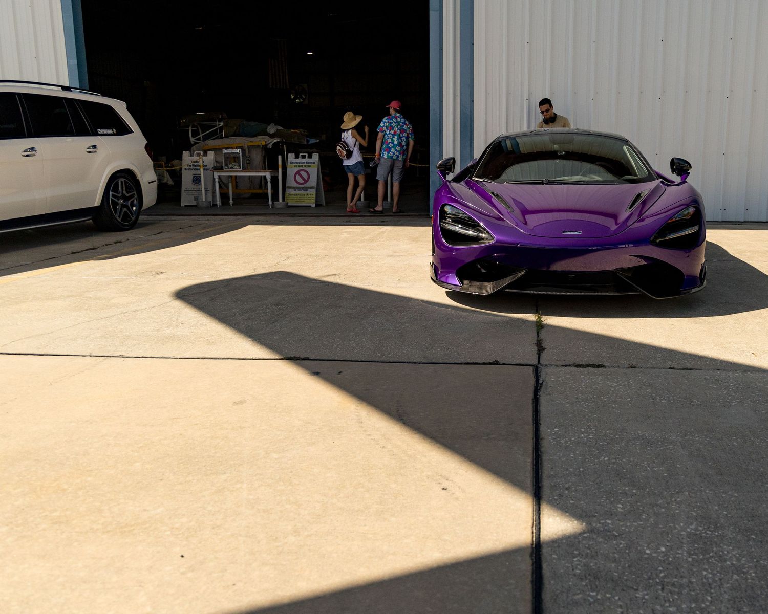 A sleek purple McLaren sports car parked in front of a white building with dramatic shadows cast on the concrete.