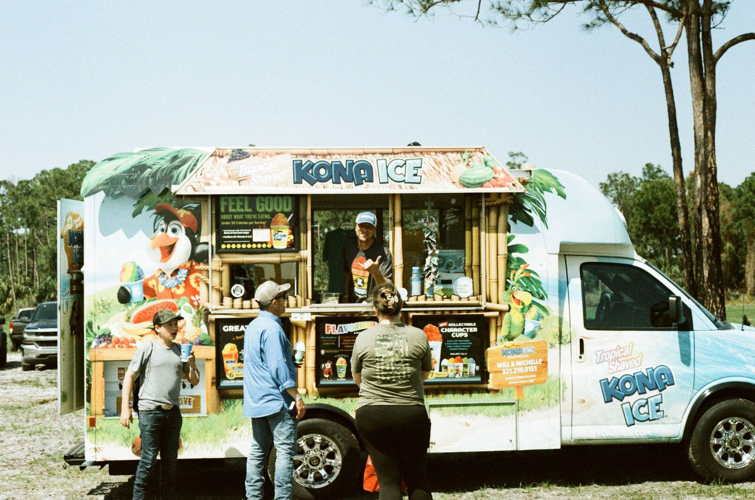 People ordering from a colorful Kona Ice shaved ice truck parked outdoors on a sunny day.