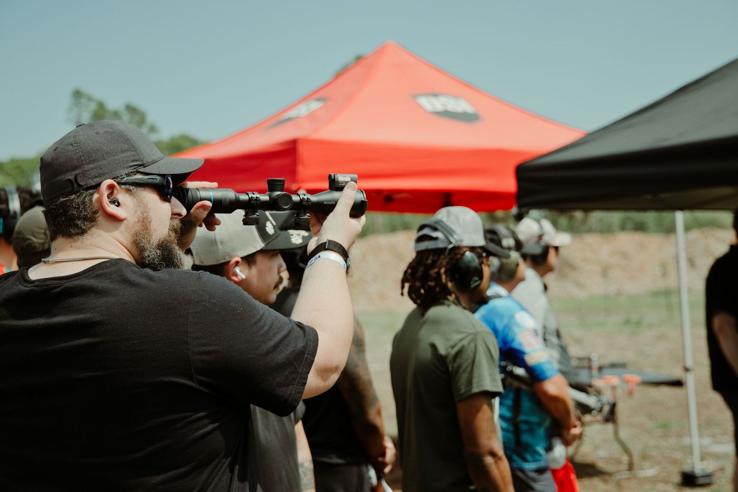 Shooters wearing ear protection aim rifles at an outdoor shooting range event with a red canopy tent.
