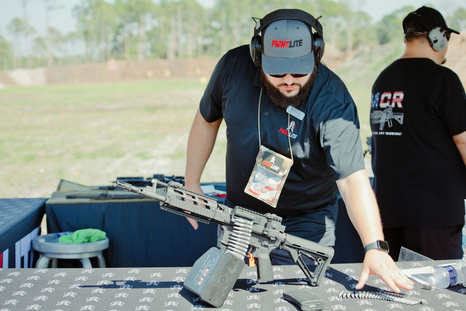 Man with beard examines tactical rifle at outdoor shooting range while wearing ear protection and a lanyard badge.