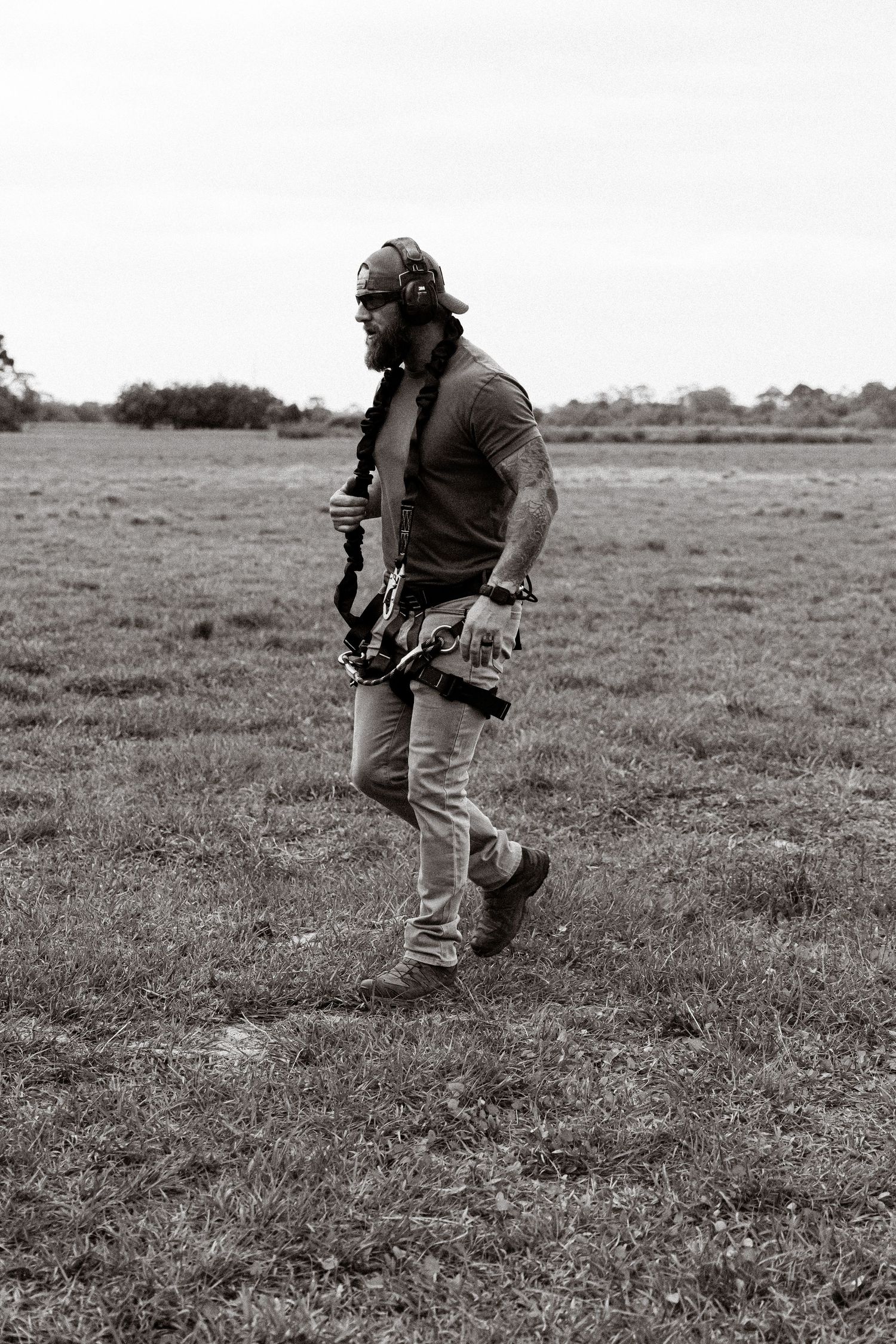 Photographer walking across an open field carrying camera equipment in black and white.