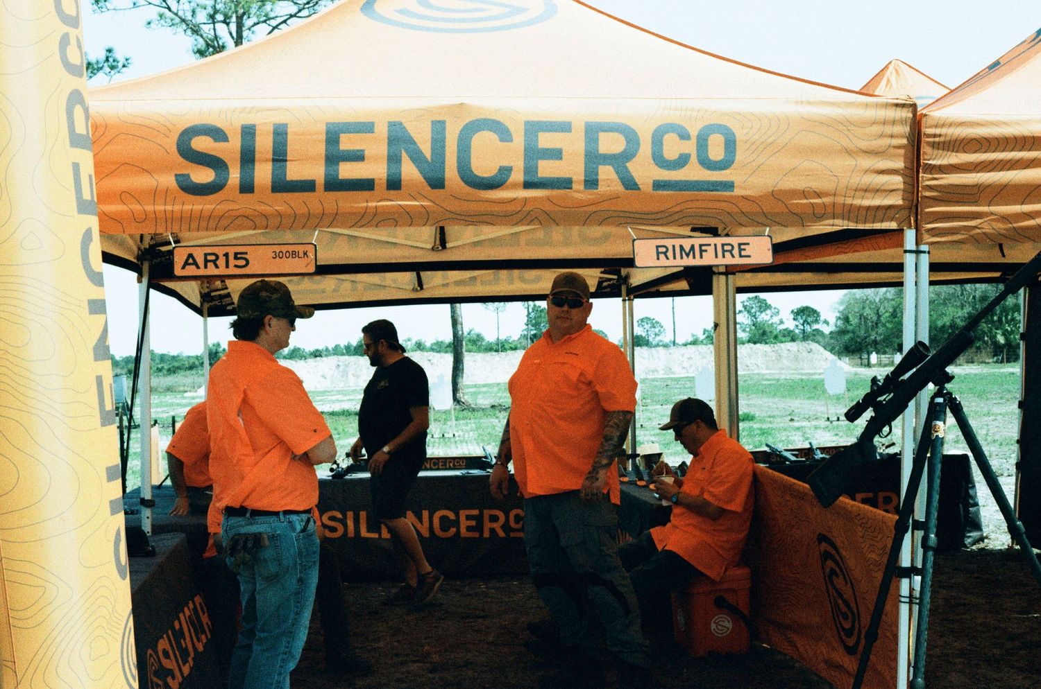 SilencerCo branded tent with staff in orange shirts showcasing AR15 and rimfire firearms at an outdoor event.