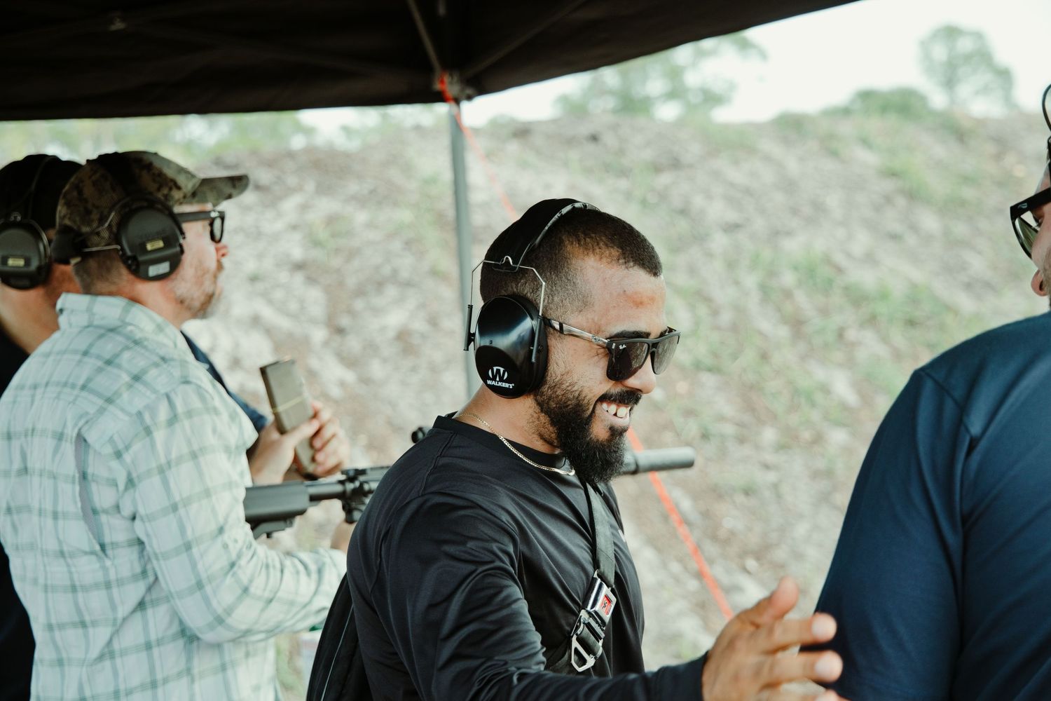 Man wearing headphones and sunglasses smiling at an outdoor shooting range.