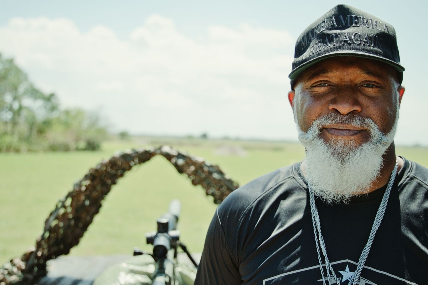 Smiling man with a white beard wearing a black cap stands outdoors near a firearm at a shooting range.