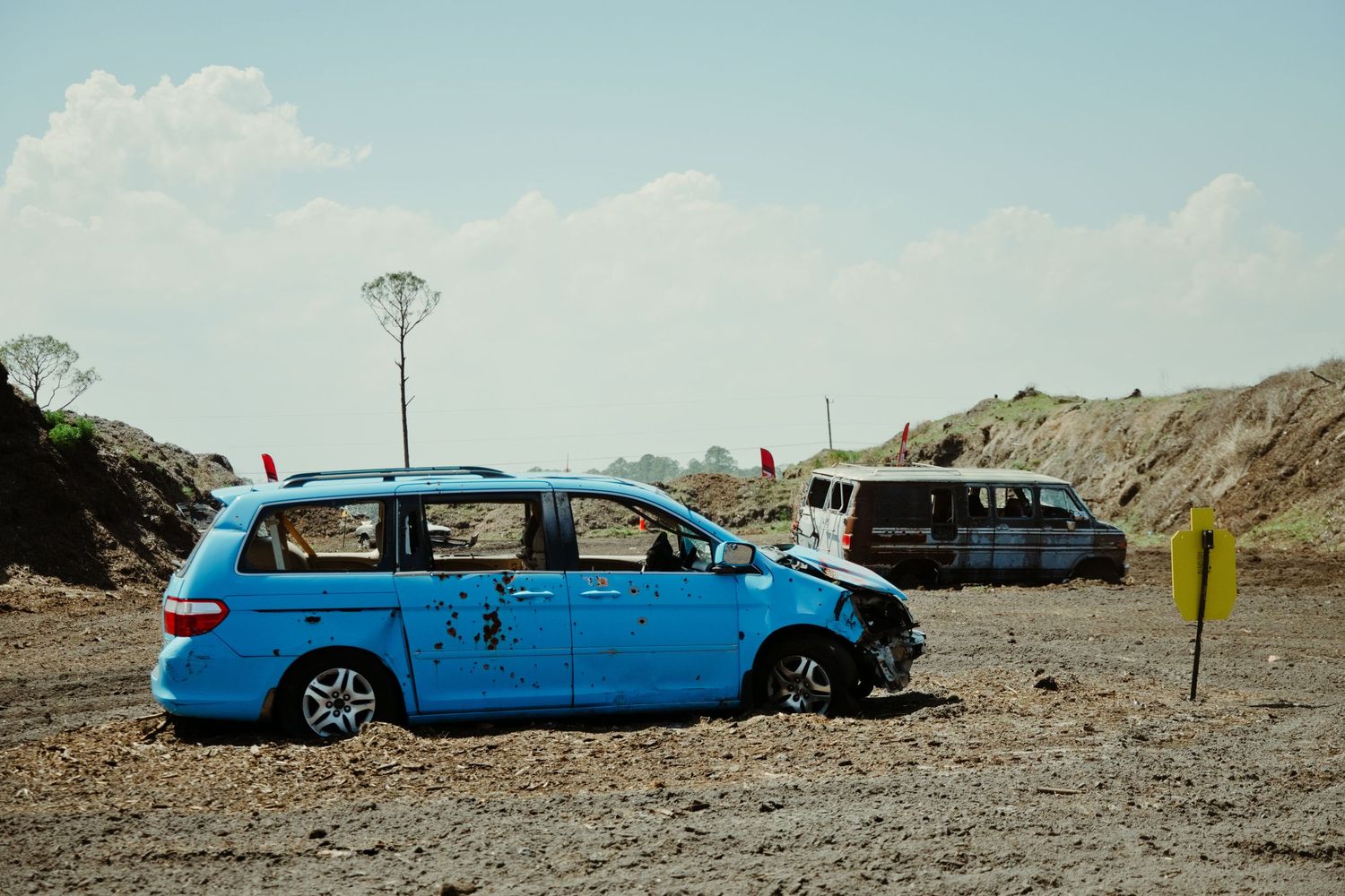 A damaged blue minivan sits abandoned on a dirt range used for shooting target practice outdoors.